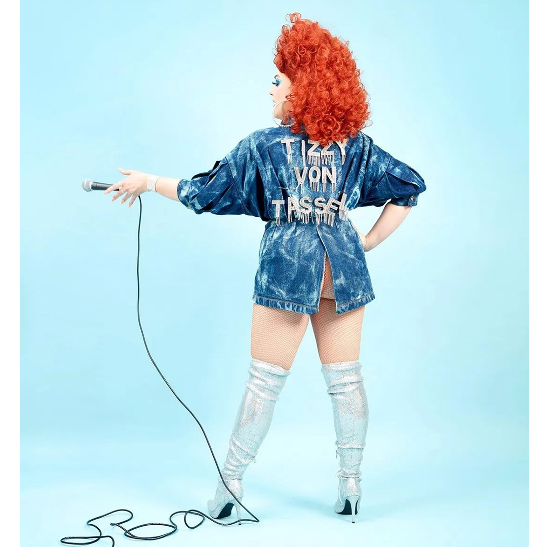 Tizzy Von Tassel, a white, orange-wig-wearing AFAB drag queen is about to perform a mic drop. She is full length, stood facing away from the camera wearing a bleached denim mini dress with 'Tizzy Von Tassel' in rhinestone letters on the back of it. You can see her butt. Background is a pale baby blue.
