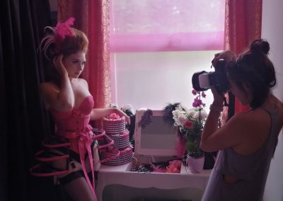 Image is mid-range and shows two women. The one on the left is white with red hair and is wearing a hot pink bra, corset and crinoline by Serefina Taylor. She has pink feathers in her hair, which is styled to look like Marie Antoinette. She is posing for the photo that is being taken by the woman on the right-a photographer mid-shot who is of east asian heritage and is wearing an oversized grey vest top. Her hair is in a messy bun on top of her head. There are pink and gold curtains in the background and they're open, revealing a pink, gauzy covering over the window. There's a dresser in the middle of the shot, with a mirror in the centre and it is draped in pearls, flowers, feathers and jewellery.